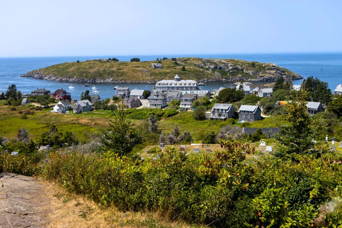 Coastal village with scattered houses, lush greenery, and Monhegan Island in the background under a clear blue sky.