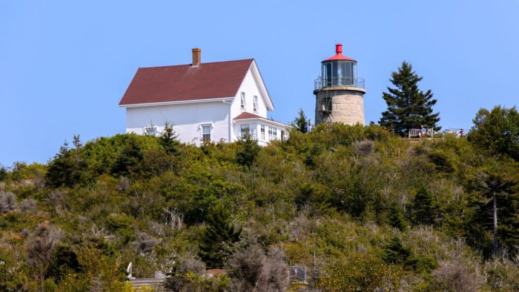 A white house and a stone lighthouse with a red roof stand atop a green, tree-covered hill under a clear blue sky.