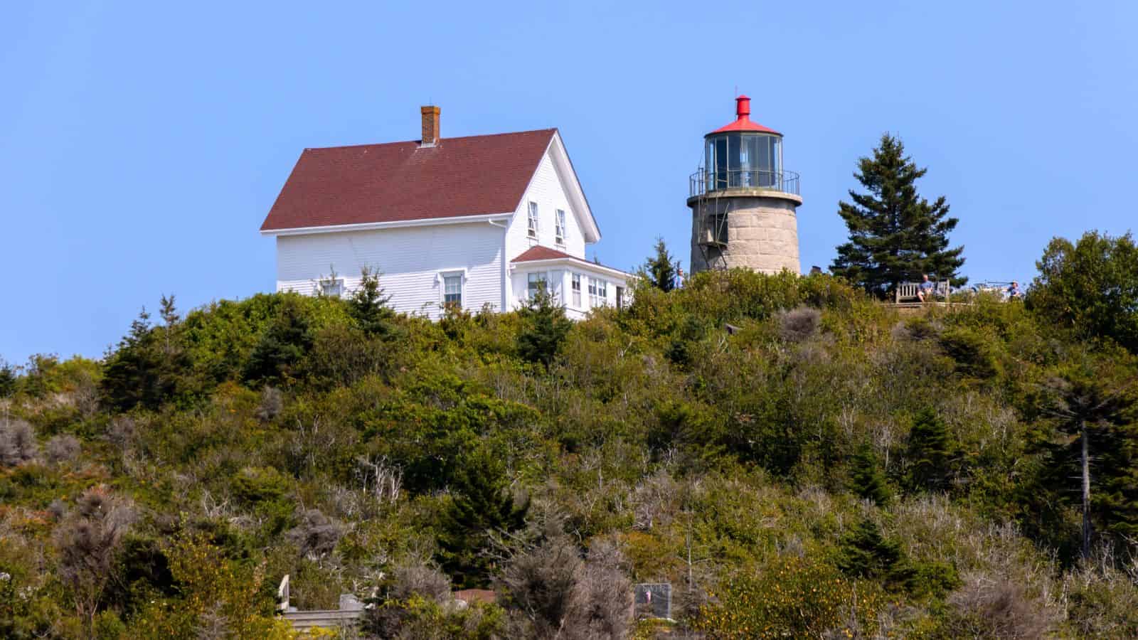 A white house and a stone lighthouse with a red roof stand atop a green, tree-covered hill under a clear blue sky.