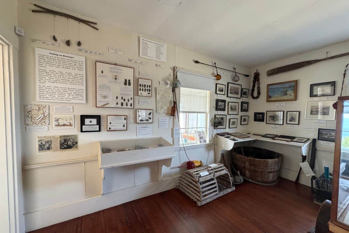 Small museum exhibit room on Monhegan Island featuring fishing artifacts, tools, framed photos, informational panels, a lobster trap, and a large wooden tub displayed against white walls and a wooden floor.