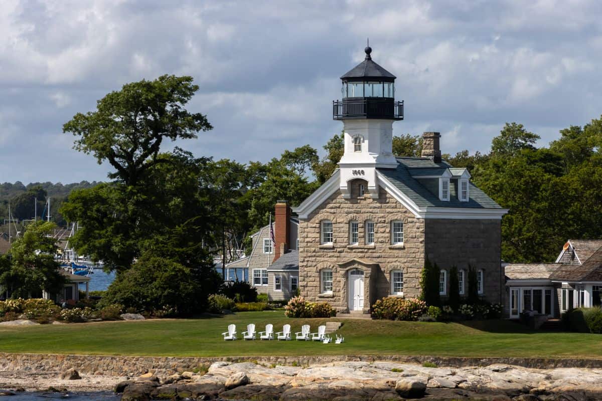 A stone lighthouse with a black lantern and white trim sits on a grassy lawn with white chairs, near water, surrounded by trees and a cloudy sky.