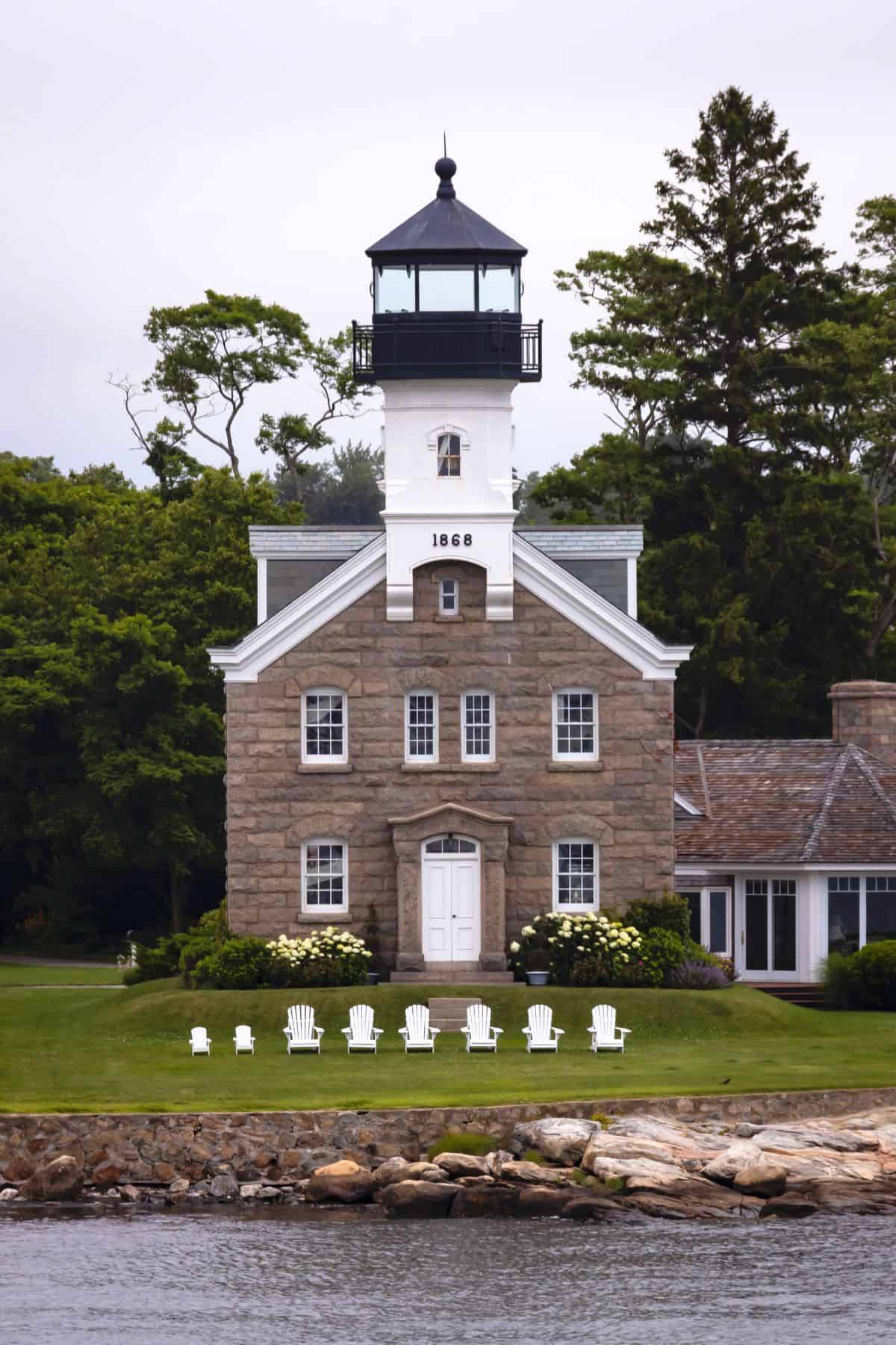 Stone lighthouse built in 1868 with white trim, surrounded by greenery, and a row of white Adirondack chairs on the lawn facing the water.