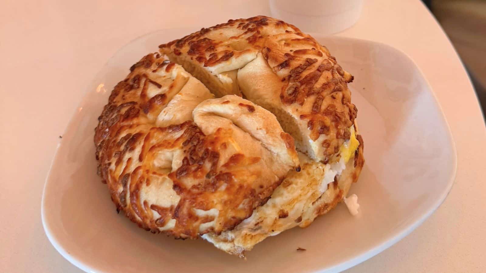 A cheese-covered bagel sliced into quarters sits on a white plate atop a light-colored table.