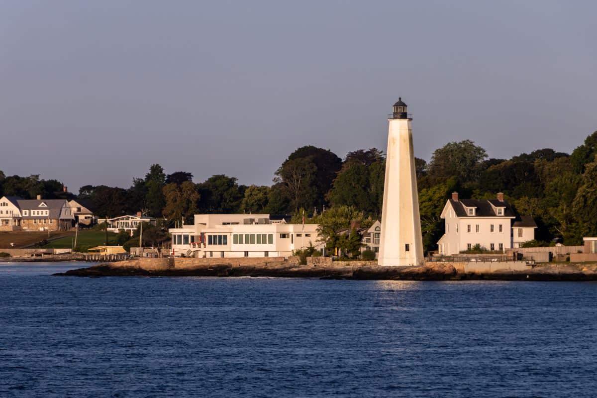 A white lighthouse stands near houses along a rocky shoreline with calm blue water in the foreground and trees in the background.