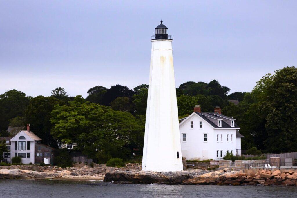 A tall white lighthouse stands on a rocky shore, with trees and houses in the background under a cloudy sky.