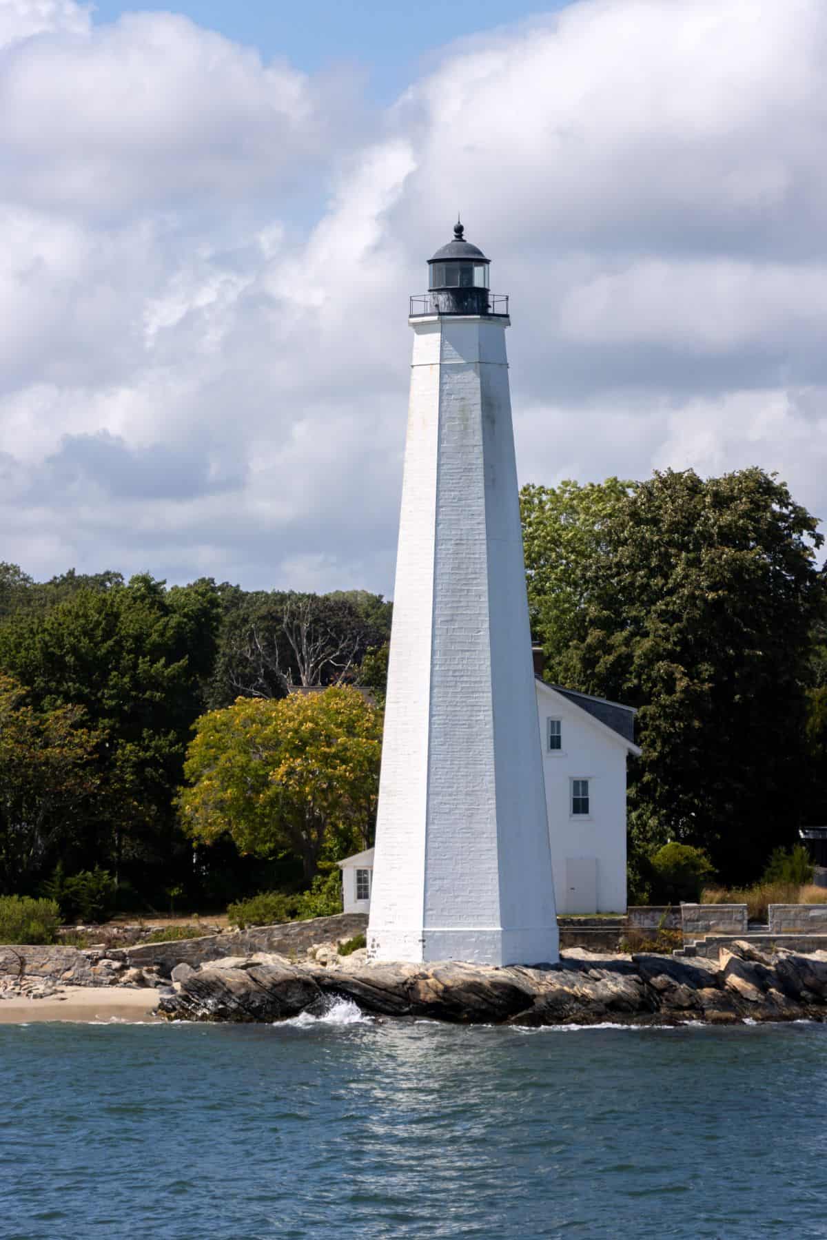 A tall white lighthouse stands on a rocky shore with a small white building behind it, surrounded by trees and water under a partly cloudy sky.