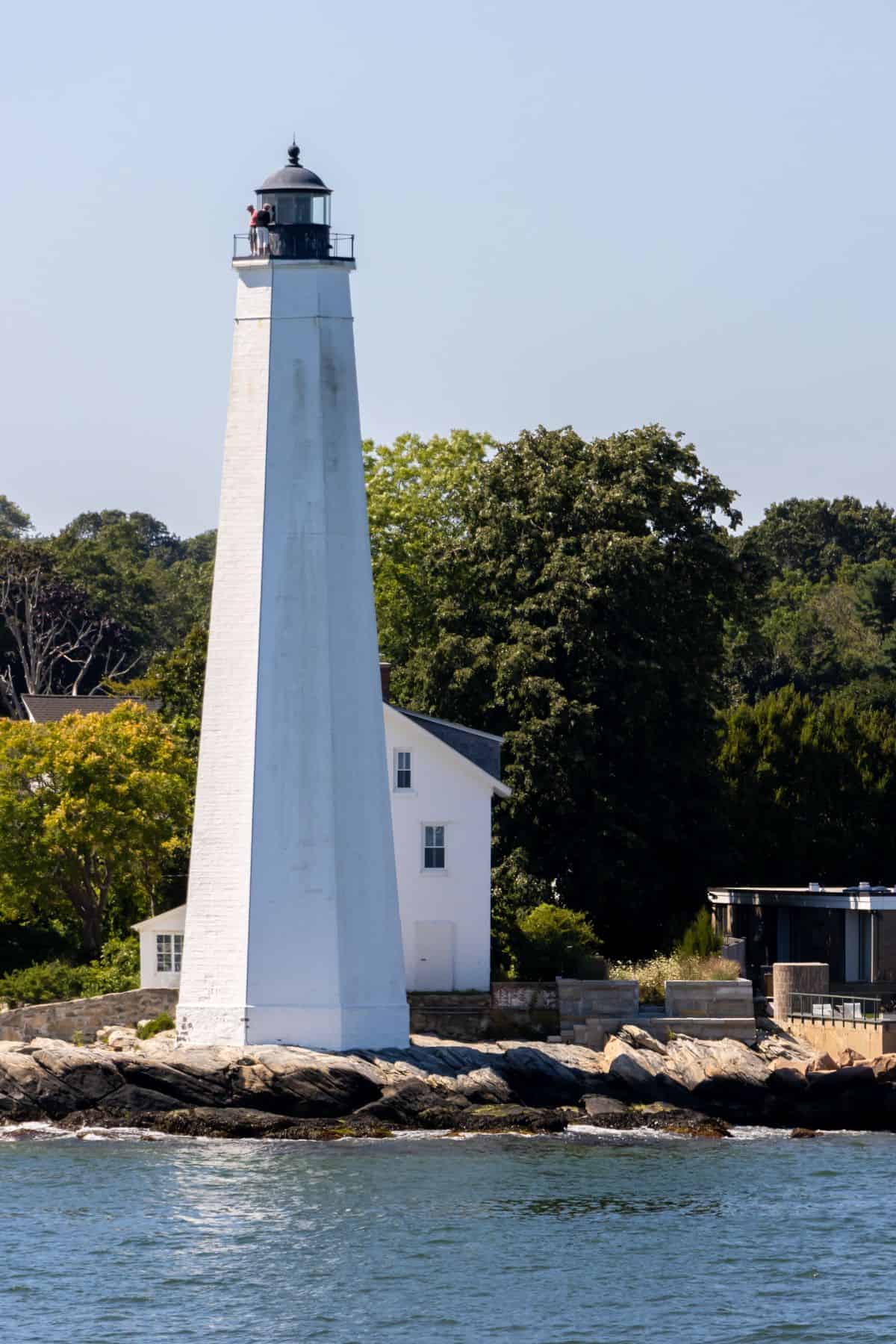 A tall white lighthouse stands on a rocky shoreline with a small white building behind it and trees in the background.