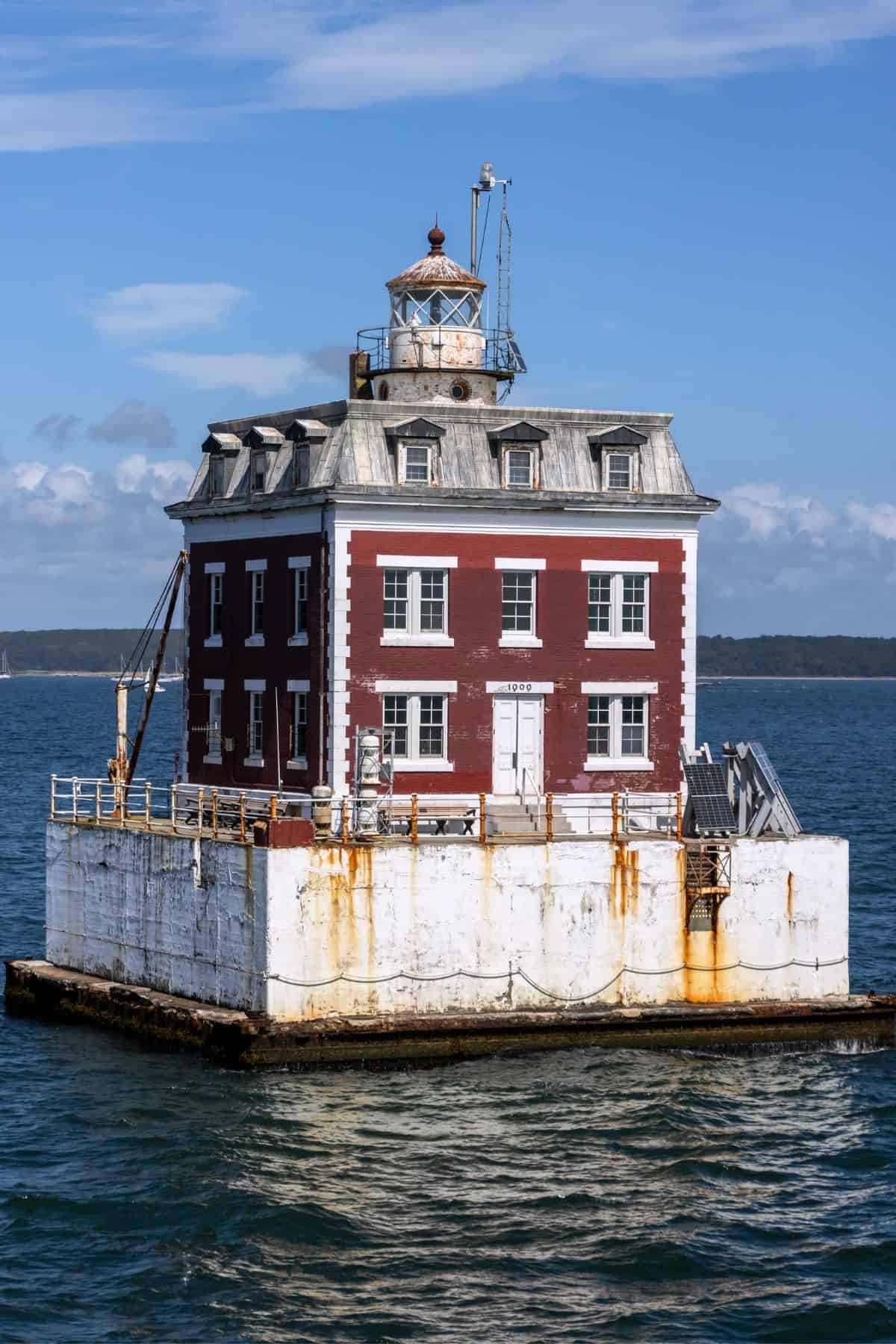 A square, red brick lighthouse with a metal roof sits on a concrete platform surrounded by water, under a partly cloudy sky.