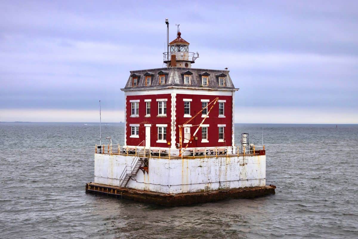Square-shaped red and white lighthouse stands on a concrete platform surrounded by water under an overcast sky.