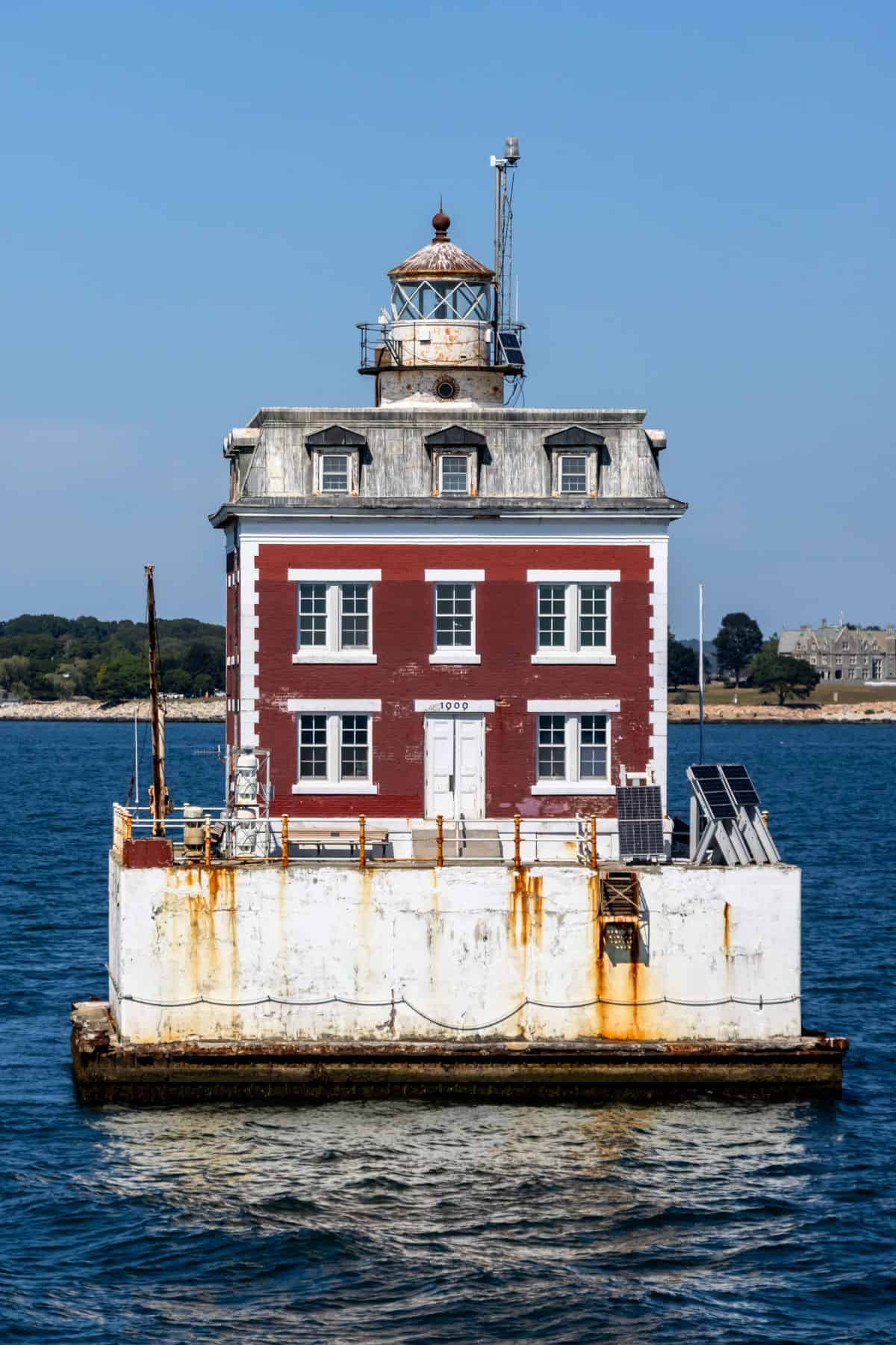 A square, red-and-white lighthouse with a rusted base sits on a concrete platform in the water near a shoreline with trees and buildings.