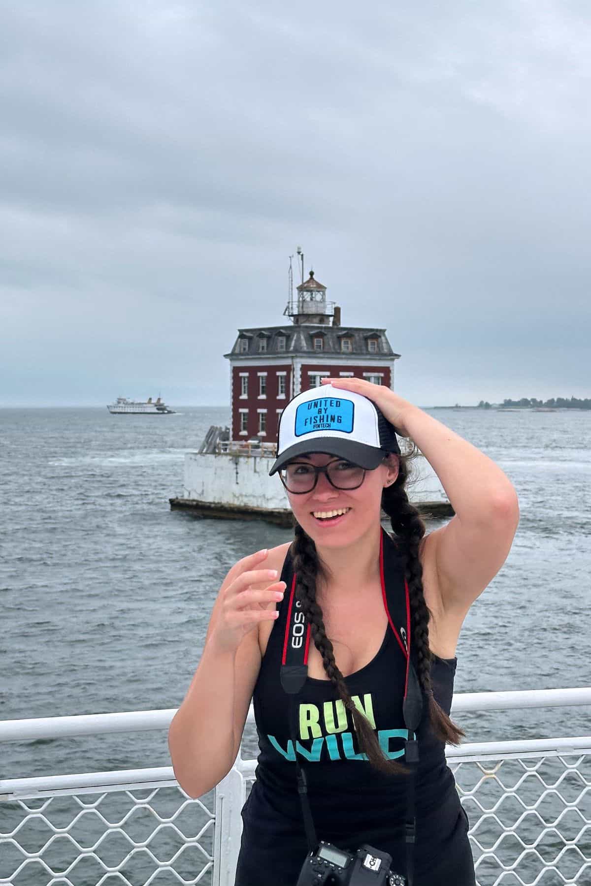 A woman wearing glasses and a cap stands on a ferry, smiling, with a camera around her neck and a lighthouse in the water behind her.