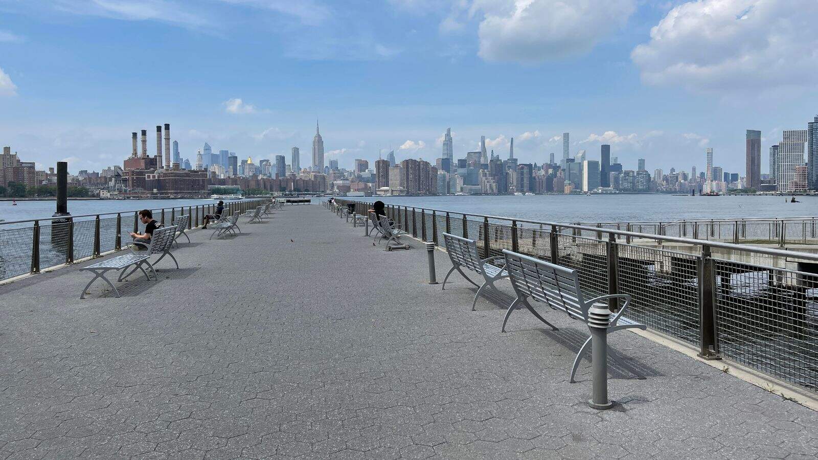 A paved waterfront pier with benches extends toward the New York City skyline under a partly cloudy sky. A few people are seated or walking along the pier.