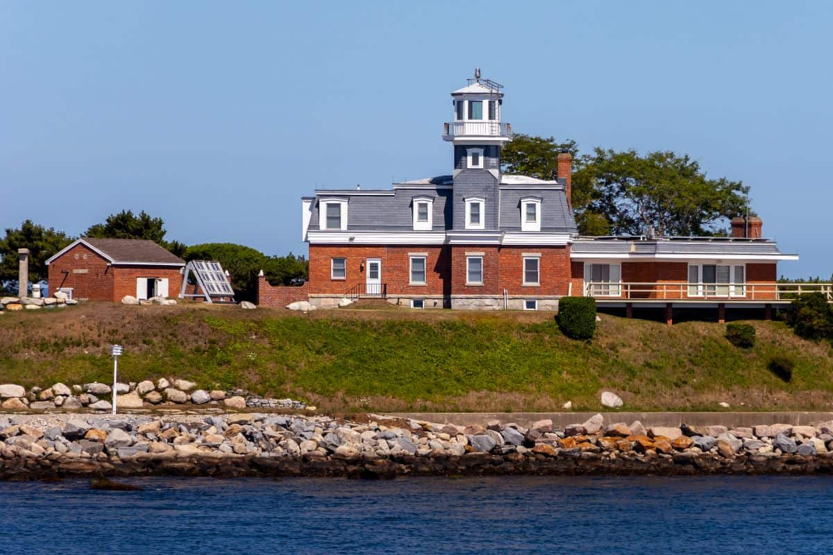 A brick lighthouse with a central tower stands on a grassy shoreline, surrounded by smaller buildings and bordered by a stone seawall next to calm blue water.