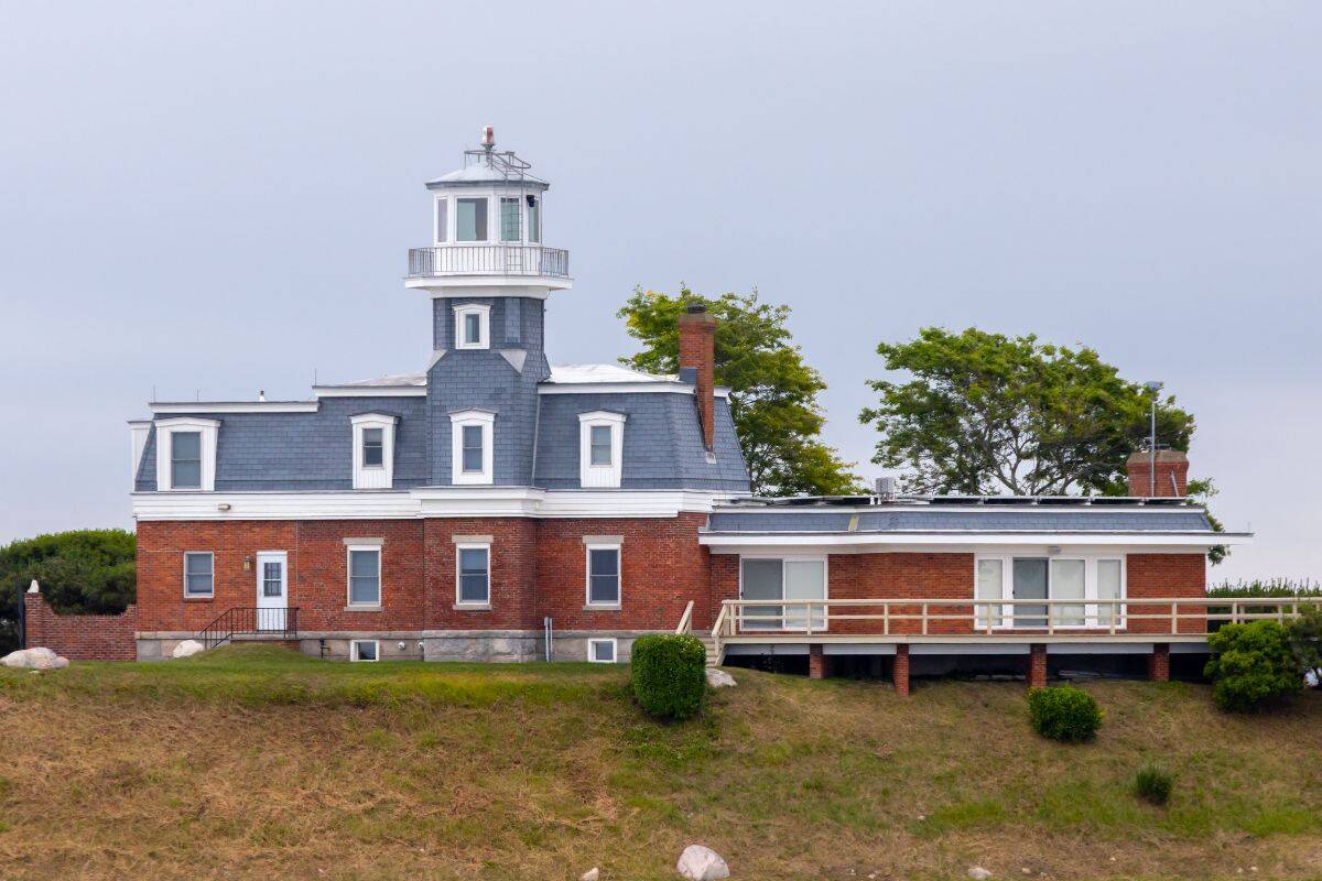 Brick lighthouse with blue-gray roof, white trim, and attached modern building sits on grassy hill with a few trees in the background under a cloudy sky—a scenic view often featured on Cross Sound Ferry lighthouse cruises.