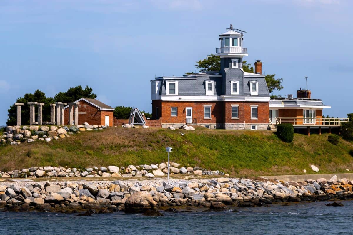 A brick lighthouse with a white lantern room stands on a rocky shoreline, surrounded by stone ruins, a small building, and greenery under a blue sky.