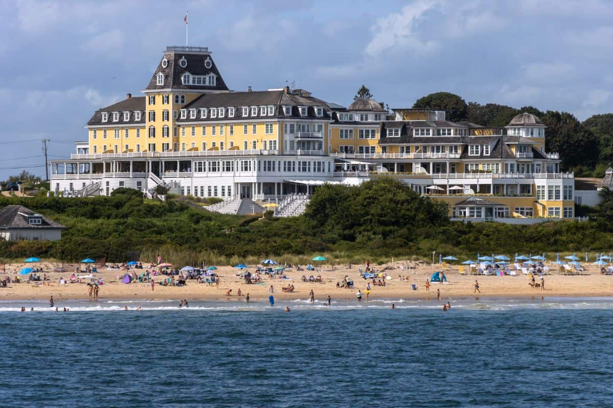 Large yellow hotel building on a hill overlooking a busy sandy beach with people sunbathing and swimming in the ocean under a partly cloudy sky.