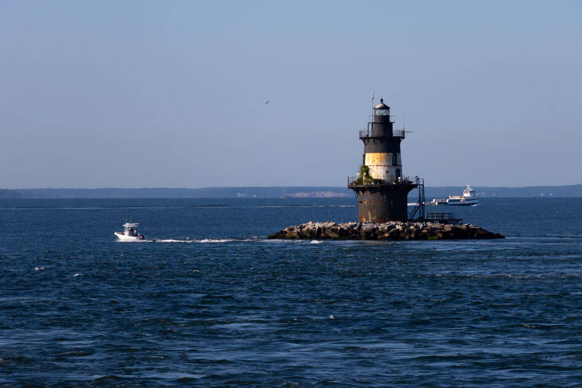 A lighthouse stands on a rocky outcrop in the ocean with a speedboat passing nearby and a ferry in the background under a clear blue sky.