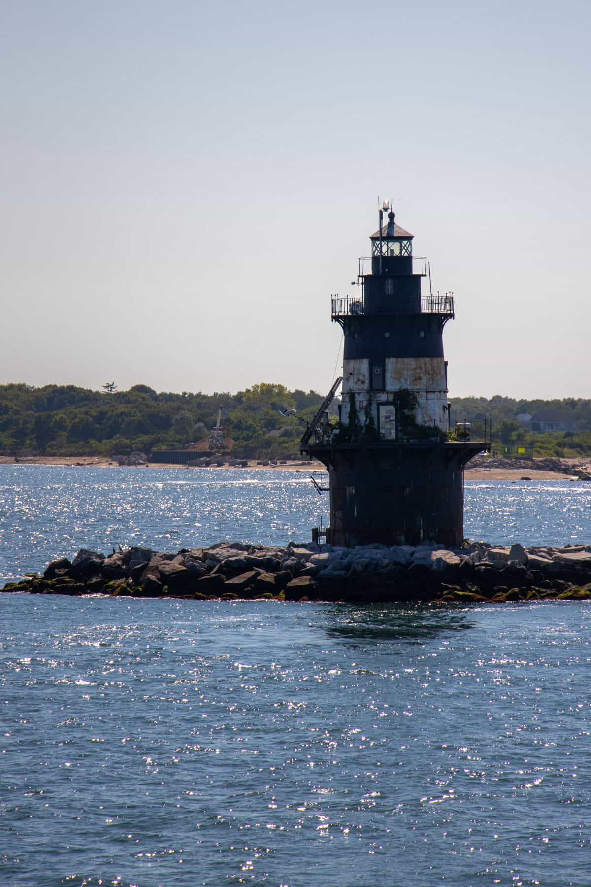 A weathered black and white lighthouse stands on a rocky breakwater surrounded by sparkling water, with a tree-lined coast visible in the background.