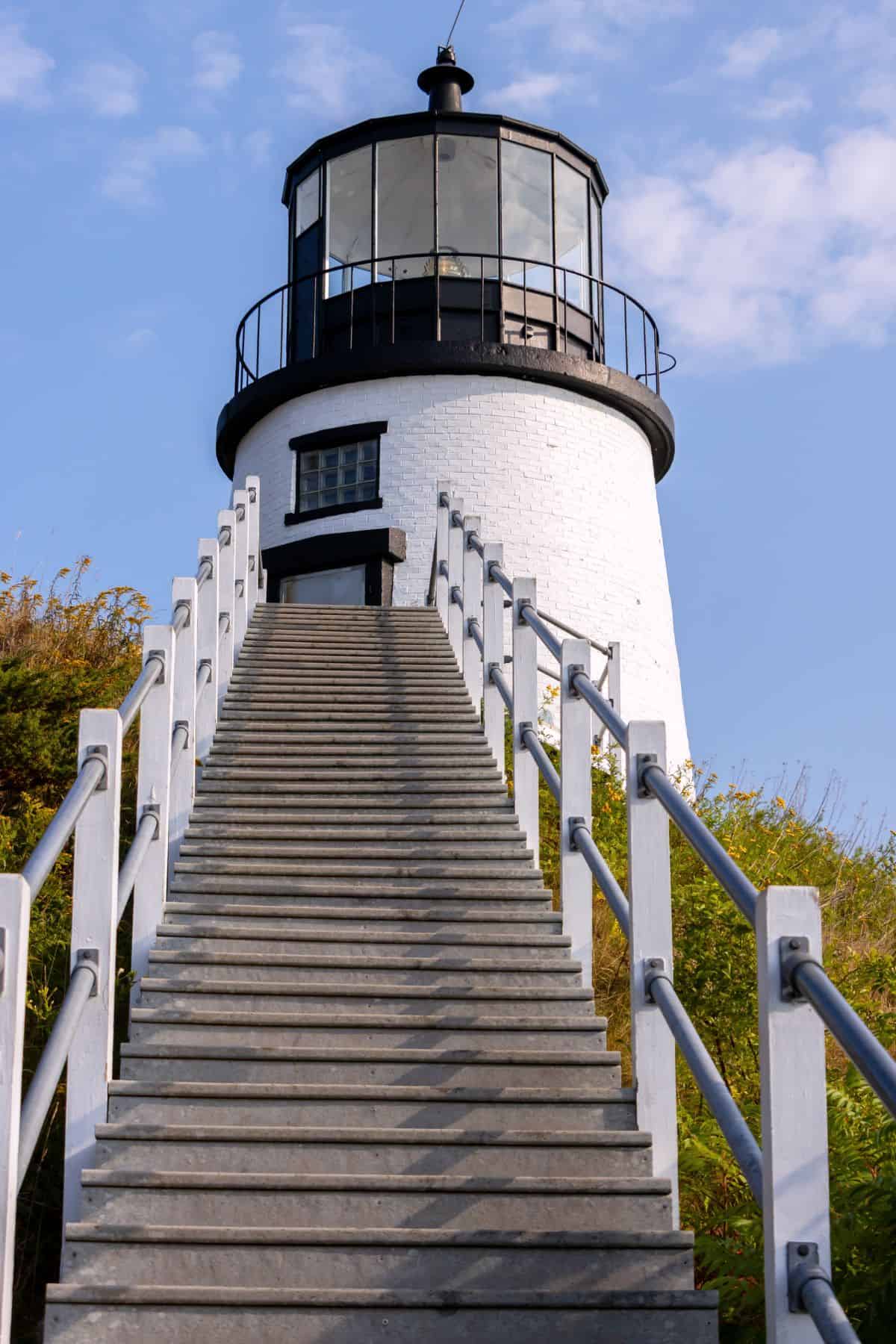 A concrete staircase leads up to a white lighthouse with a black lantern room, set against a blue sky with some clouds.