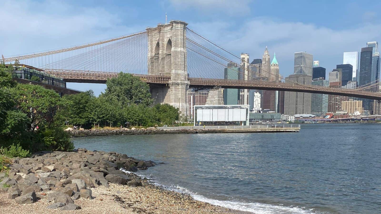 View of the Brooklyn Bridge spanning over the East River with Manhattan skyline in the background, seen from a rocky shoreline and greenery in the foreground.