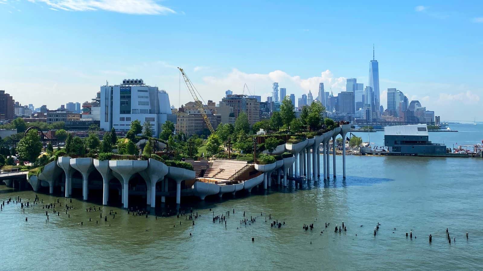 A modern park with elevated platforms and greenery sits over the water, with the New York City skyline visible in the background.