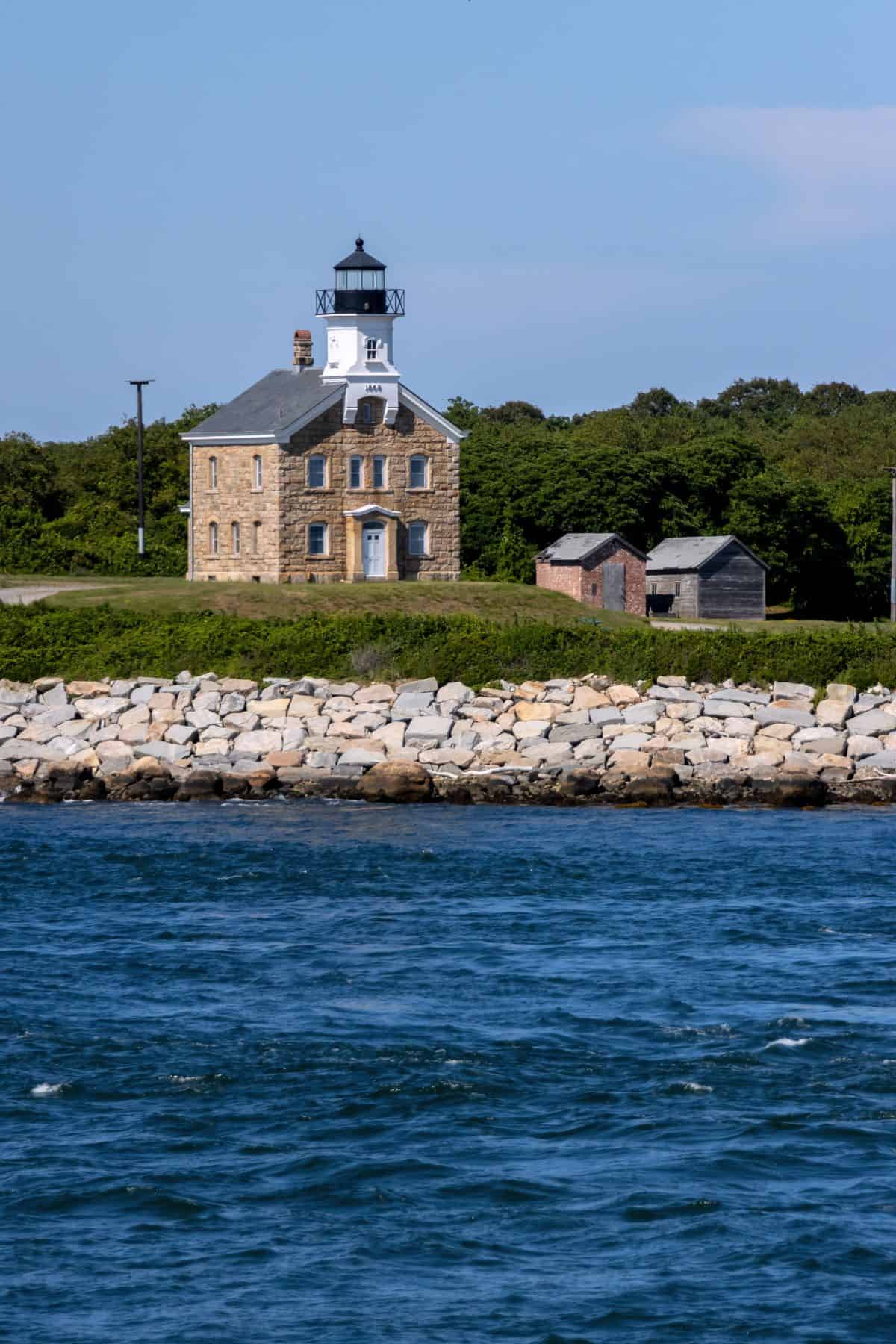 A stone lighthouse with adjacent small buildings stands near a rocky shoreline, bordered by trees and blue water under a partly cloudy sky.