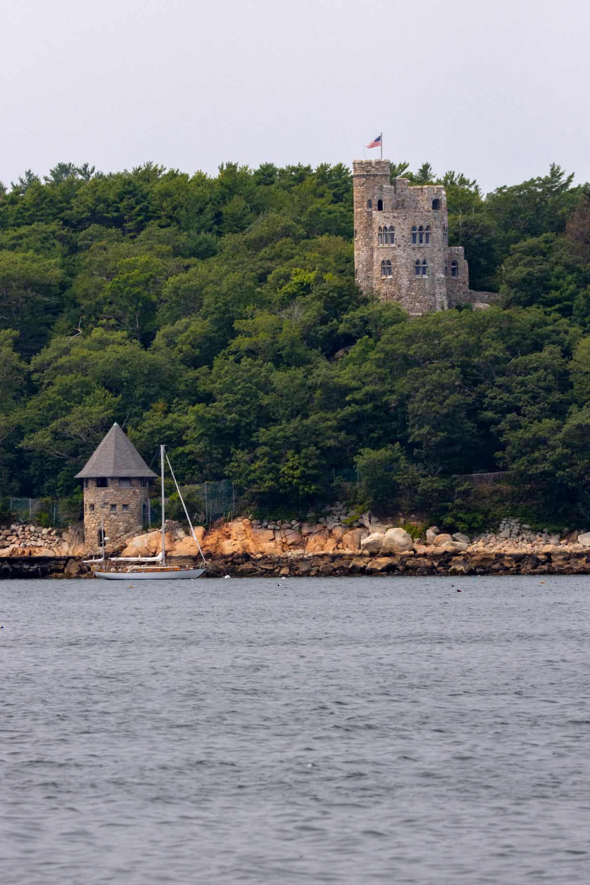 A stone tower with a flag stands on a forested hill above a rocky shoreline and a docked sailboat, with water in the foreground.