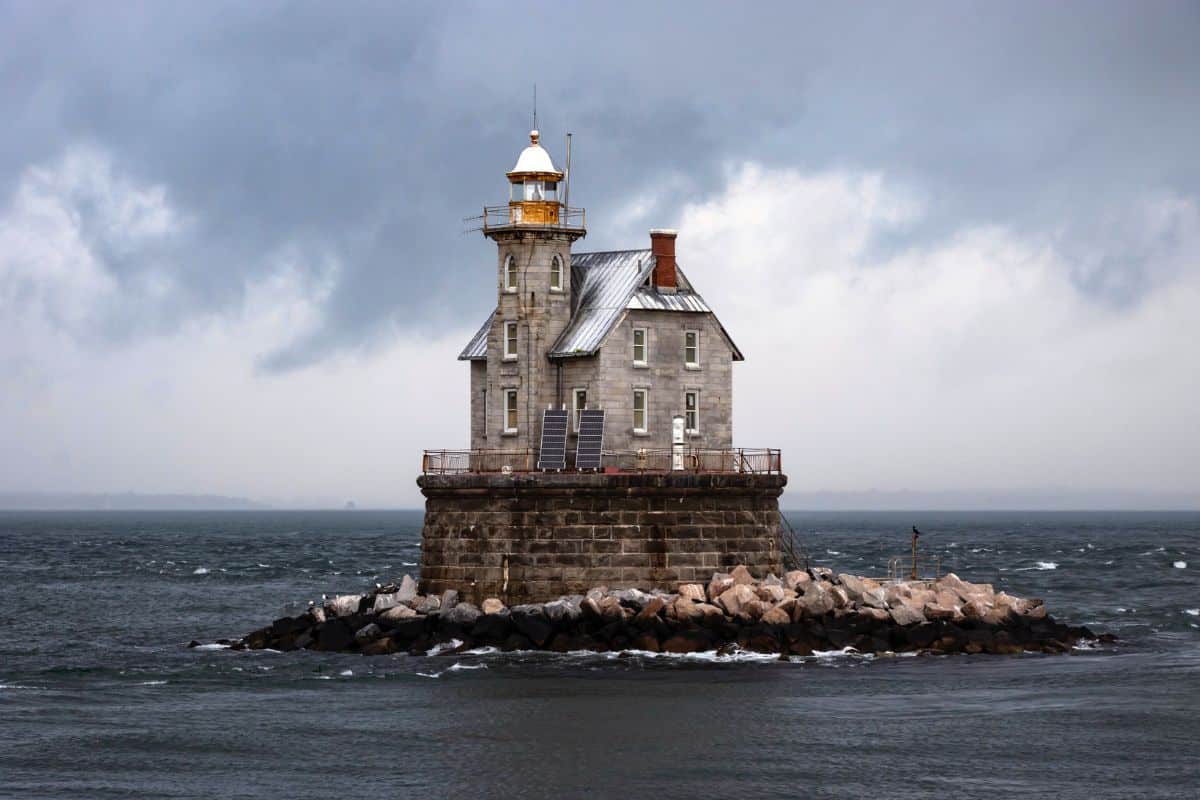A stone lighthouse with a metal roof stands on a rocky platform surrounded by choppy water under a cloudy, overcast sky.