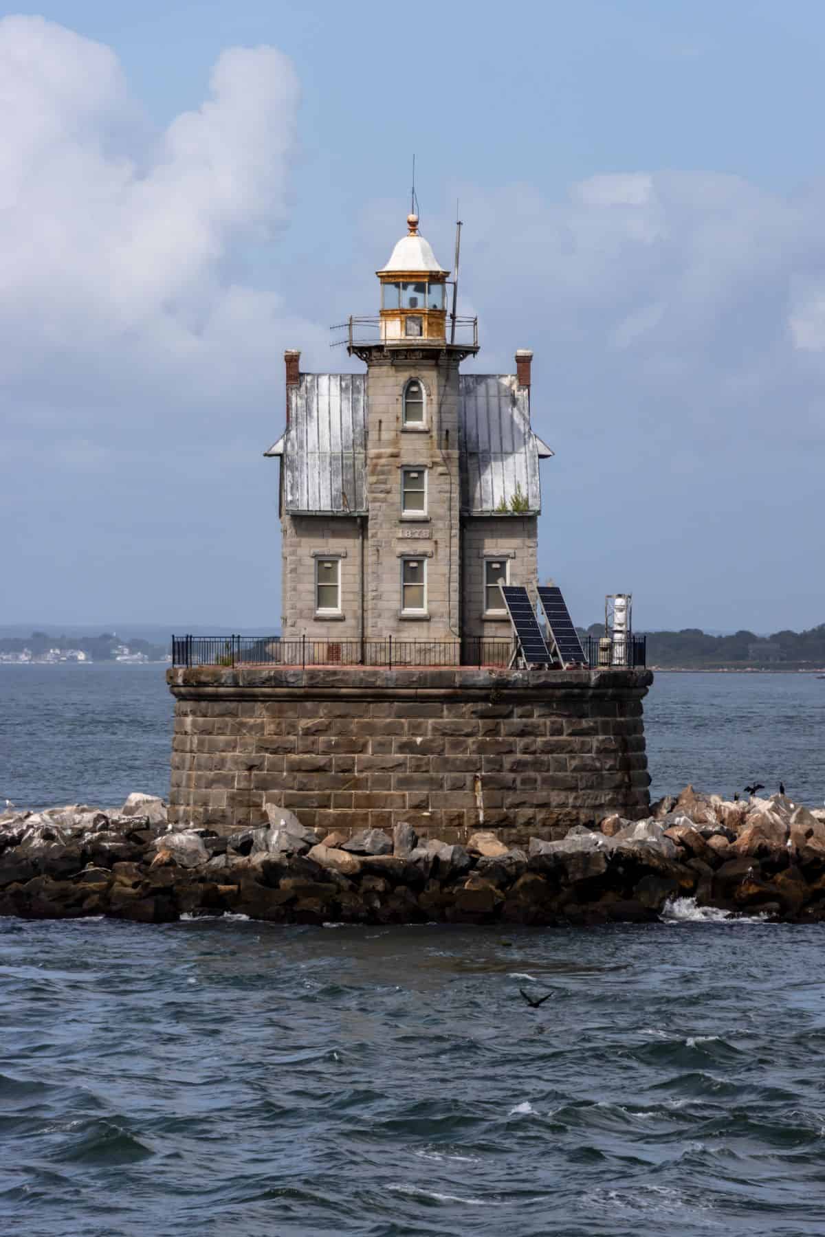 Stone lighthouse with a weathered metal roof stands on a rocky barrier in the water, surrounded by a metal fence and solar panels, with land visible in the distance.