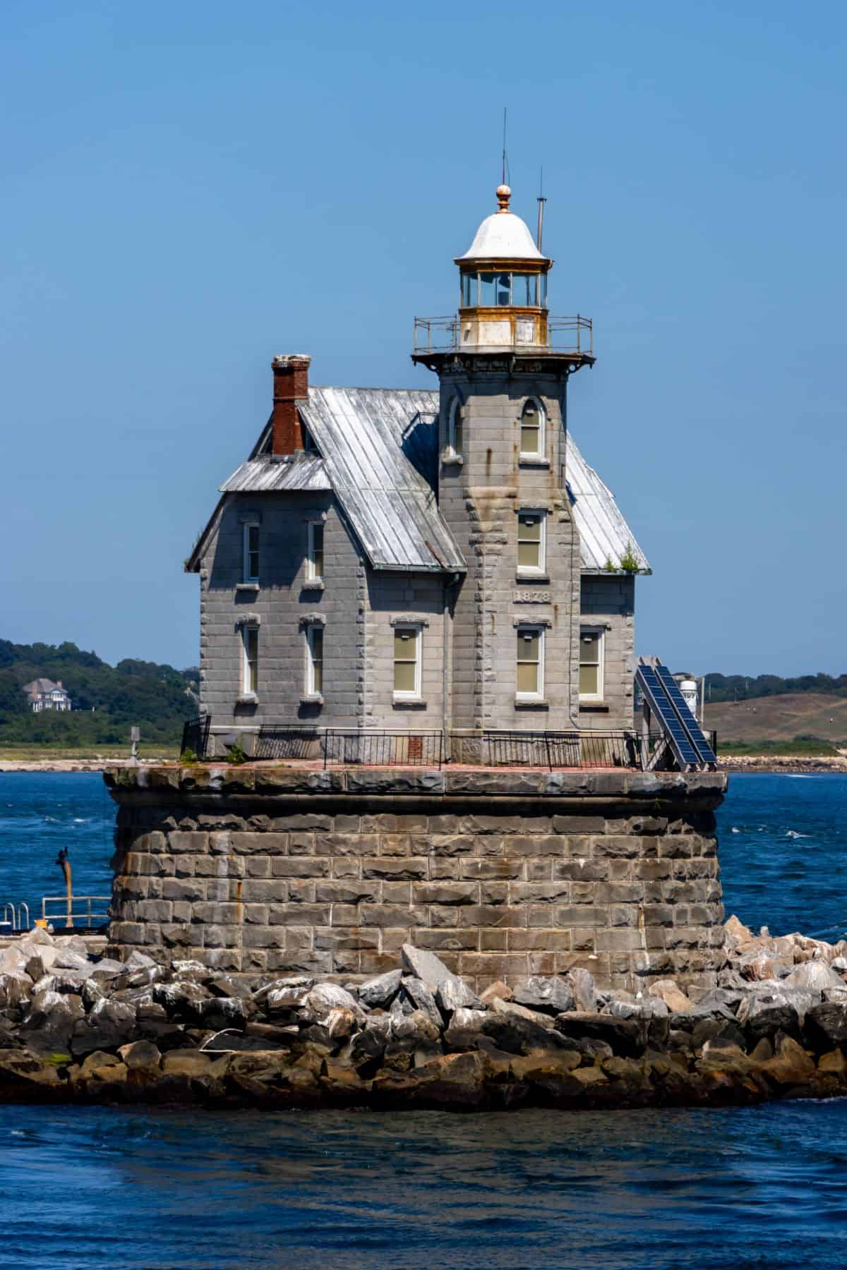 A stone lighthouse with a metal roof stands on a rocky base surrounded by water, with distant land visible in the background under a clear blue sky.