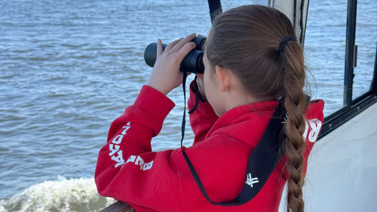 A girl with a long braid in a red hoodie looks through binoculars while standing on a boat next to a body of water.