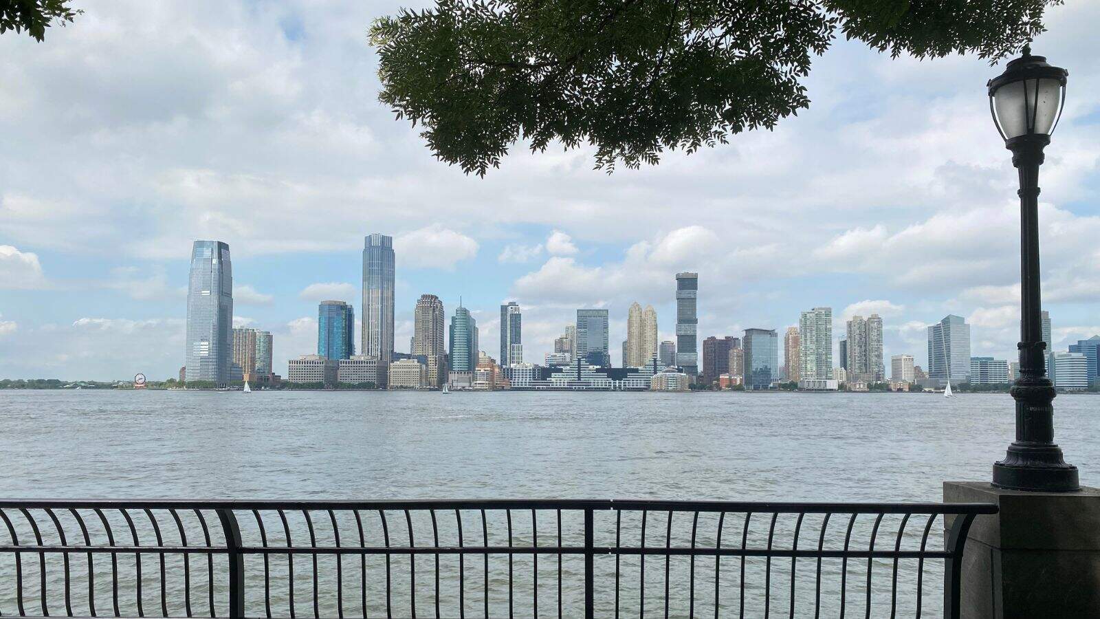 View of a city skyline across a river with modern skyscrapers, a cloudy sky, tree branches above, and a black metal fence and streetlamp in the foreground.