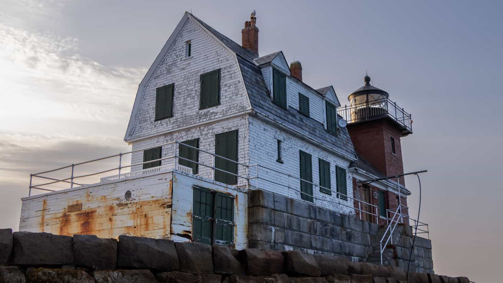 A weathered white lighthouse keeper's house with green shutters stands on a stone foundation next to a red brick lighthouse tower under a cloudy sky.