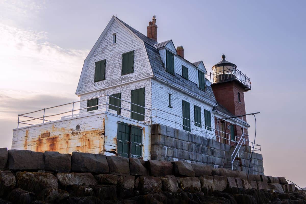 A weathered, white wooden lighthouse with boarded windows and a rusty section sits on a stone foundation by the water under a partly cloudy sky.