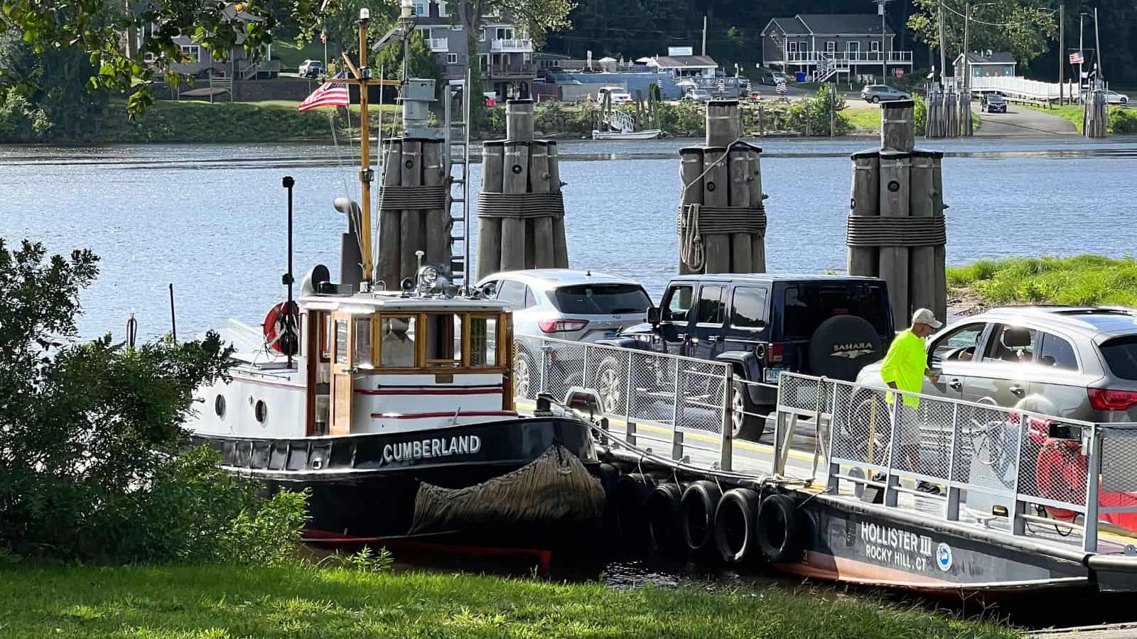 A small ferry named Cumberland transports cars and passengers across a river; vehicles and people are visible on board, with buildings and trees in the background.