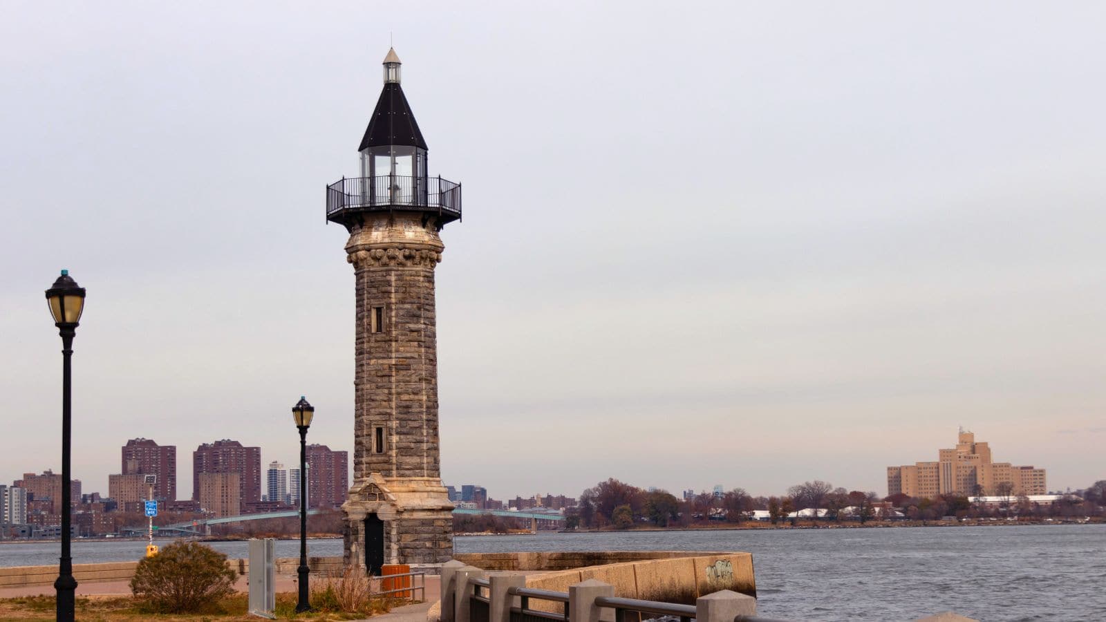 A stone lighthouse stands by the water with a city skyline and buildings in the background under an overcast sky.