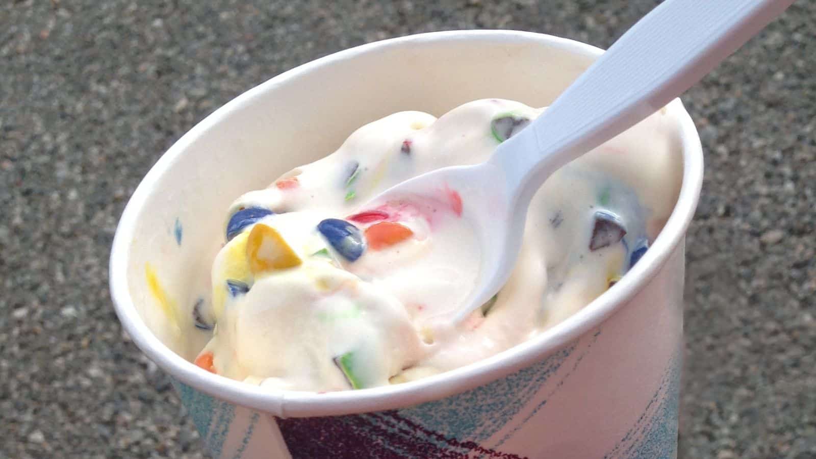 A cup of vanilla soft serve ice cream mixed with colorful candy pieces, served with a white plastic spoon.