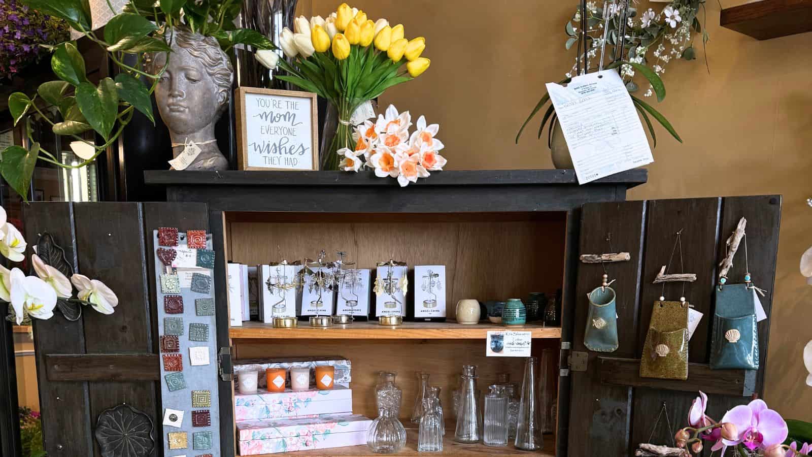 A wooden cabinet with shelves displays glass bottles, decorative boxes, greeting cards, ceramic items, and vases, with flowers and a bust on top. A sign reads, "You are the mom everyone wishes they had.