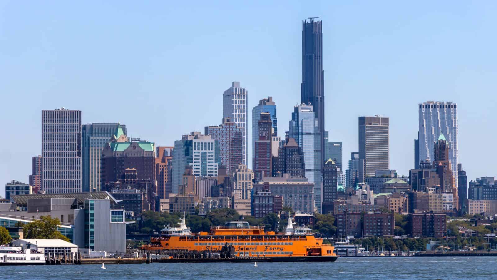 A bright orange Staten Island Ferry crosses the river with the Manhattan skyline visible in the background on a clear day.