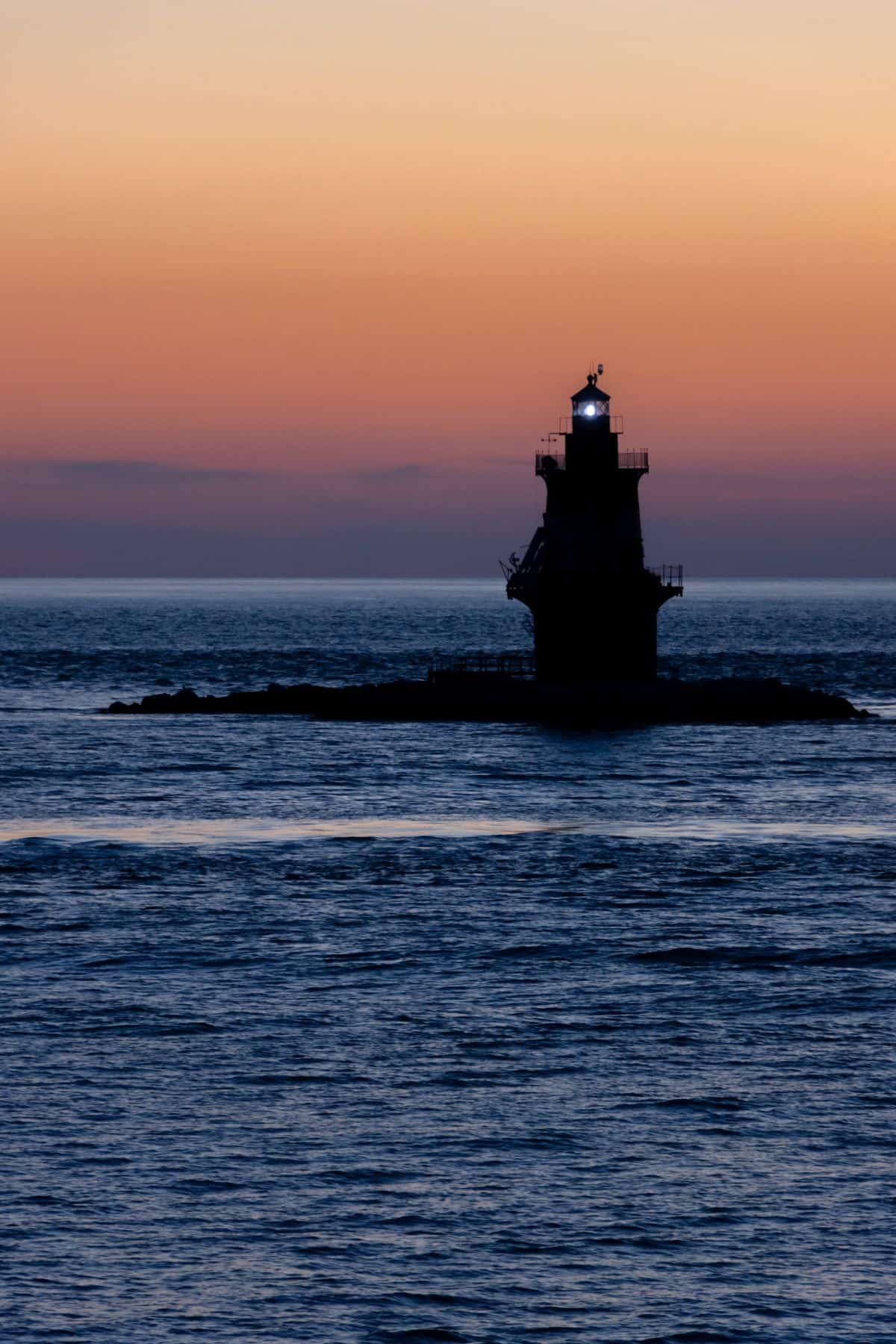 A lighthouse with its light on stands on a small rocky island, silhouetted against an orange and blue sunset sky over the calm sea.