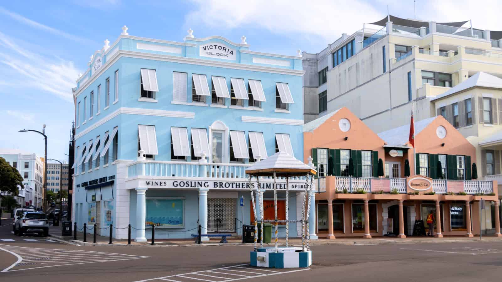 A street corner with pastel-colored buildings, including the blue Victoria Block and pink stores, and a decorative well in the foreground under a partly cloudy sky.