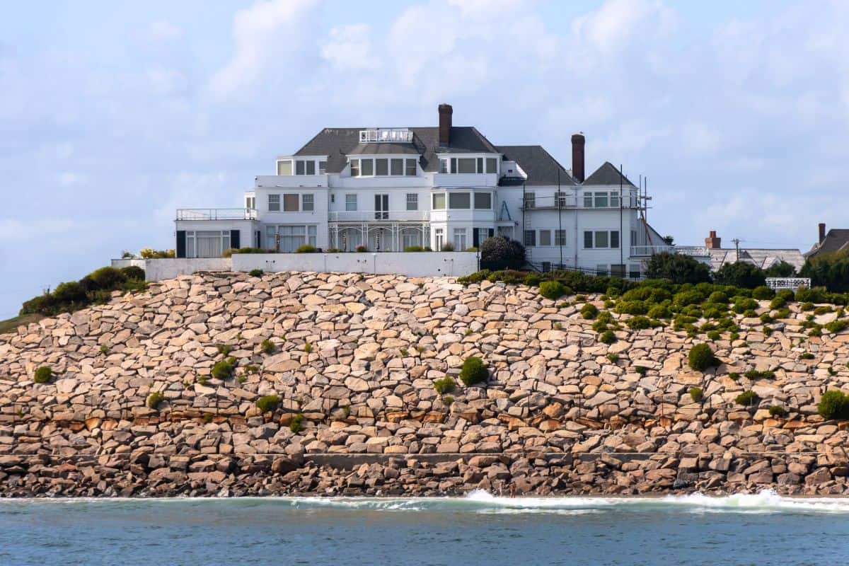 Large white house with multiple chimneys sits atop a rocky hillside overlooking the water, under a partly cloudy sky.