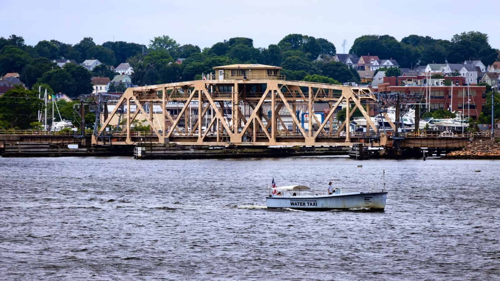 A water taxi crosses a river with a metal drawbridge and houses visible in the background under a cloudy sky.