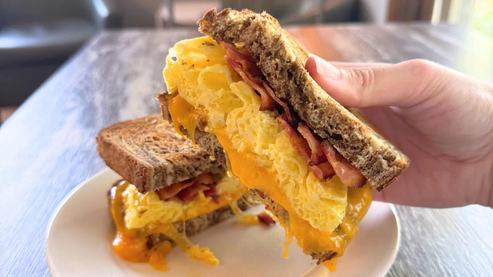 A hand holds a breakfast sandwich with scrambled eggs, bacon, and melted cheddar cheese between two slices of toasted whole grain bread on a white plate.