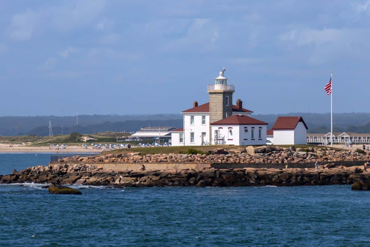 A lighthouse and adjacent buildings sit on a rocky shoreline next to the ocean, with an American flag and beach visible in the background.