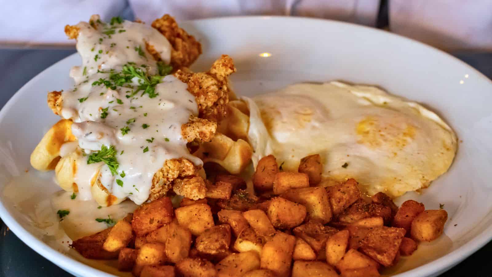 A plate with fried chicken on waffles topped with white gravy, two sunny-side up eggs, and seasoned diced potatoes—this hearty dish is just one of the mouthwatering options you’ll find at top breakfast places in Connecticut.