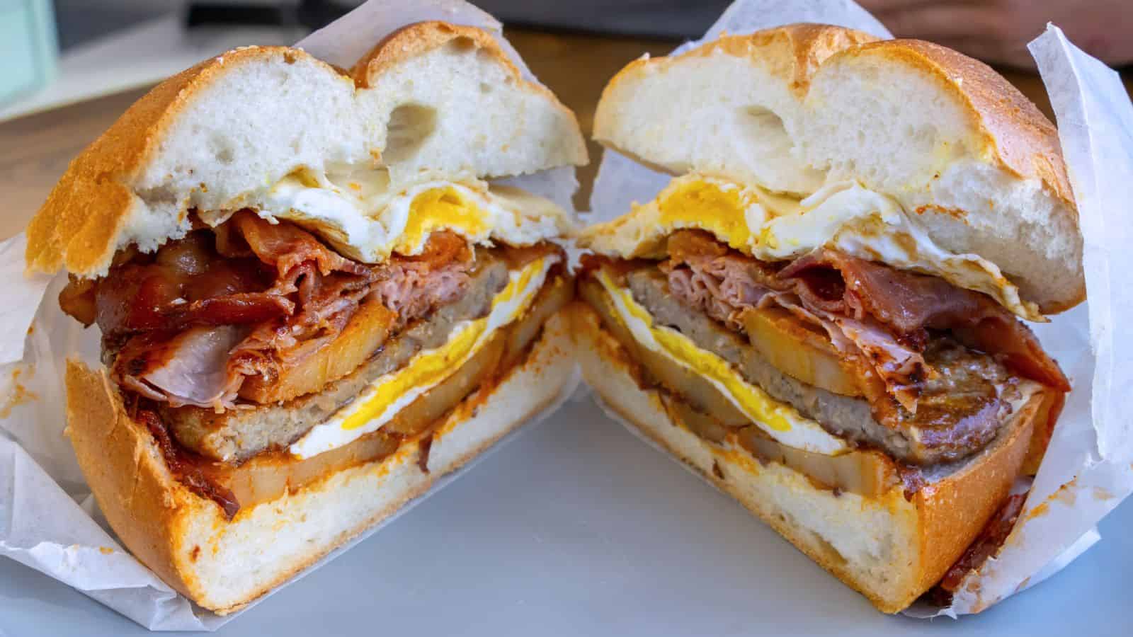 A breakfast sandwich cut in half, showing layers of egg, bacon, sausage, cheese, and possibly potato on a round bun—just like the delicious options you'd find at top breakfast places in Connecticut.