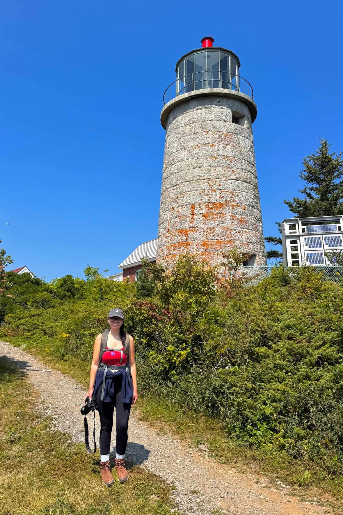 A woman in athletic clothing stands on a dirt path holding a camera, with Monhegan Island’s old stone lighthouse and lush greenery in the background under a clear blue sky.