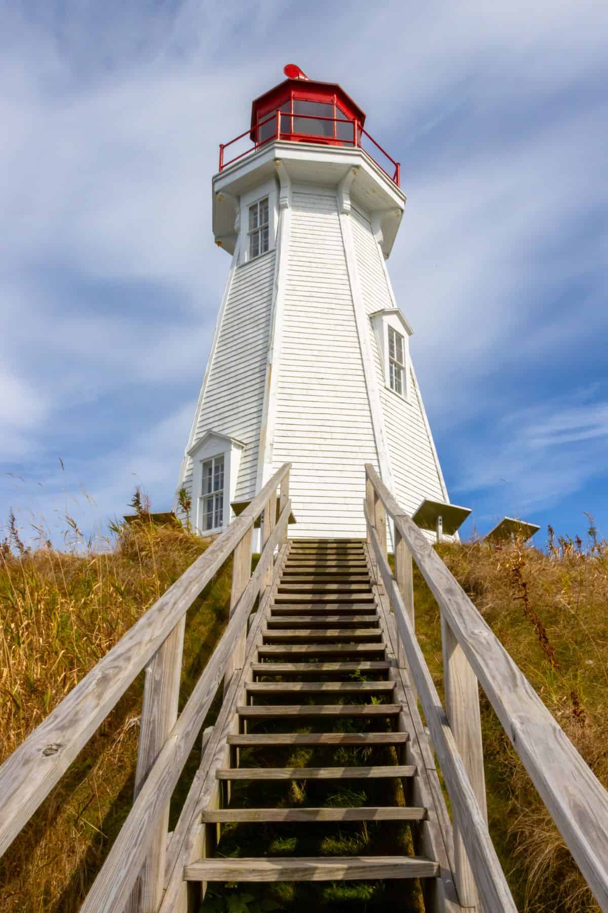 A white lighthouse with a red top stands at the top of a wooden staircase, surrounded by tall grass under a partly cloudy sky.