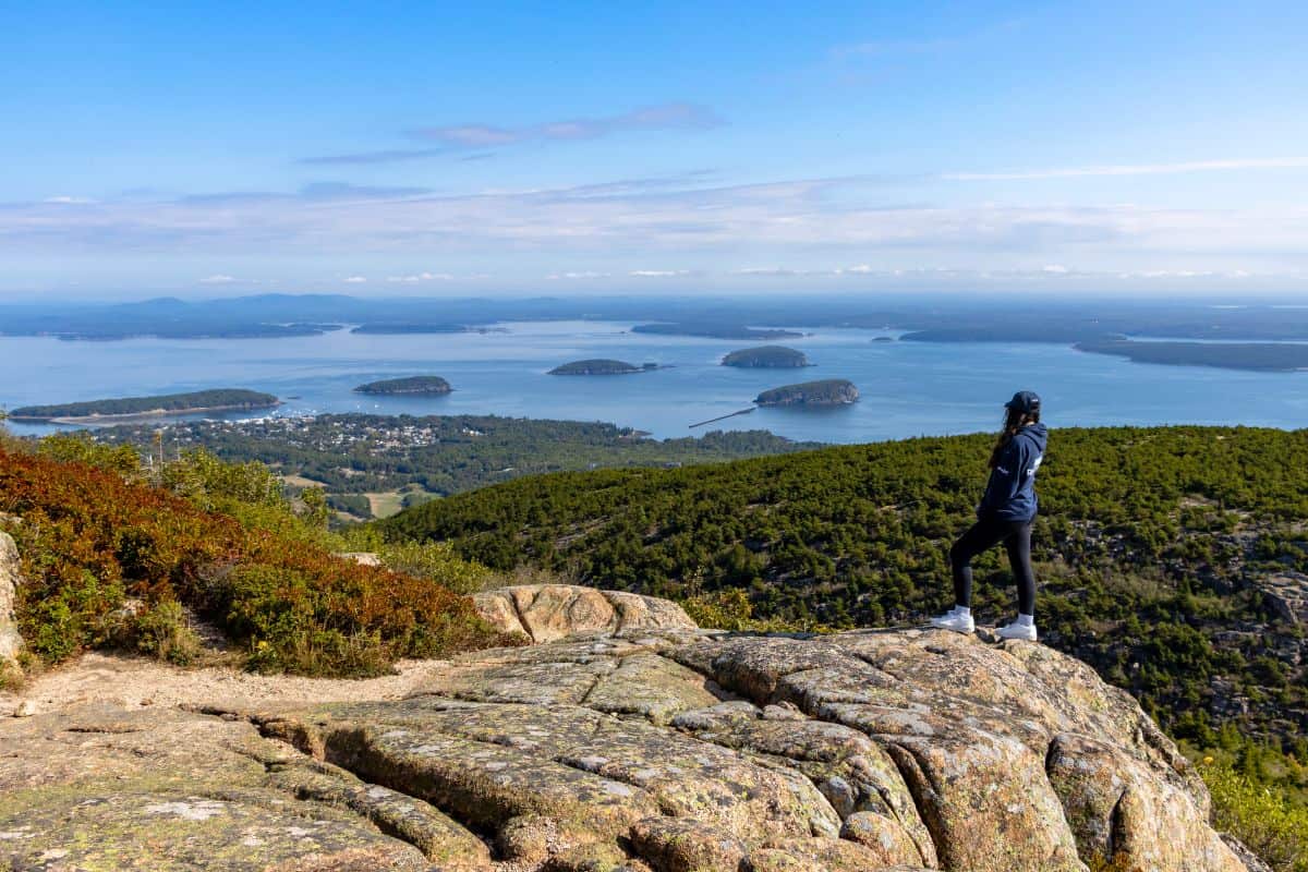 A person stands on a rocky ledge overlooking a forested landscape and several islands in a body of water under a clear sky.